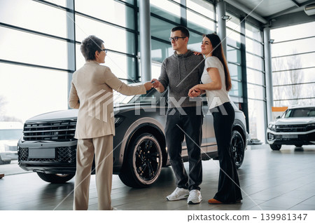 Couple is shaking hands with a saleswoman in a modern car dealership, showcasing a successful vehicle purchase and a welcoming atmosphere Couple is shaking hands with a saleswoman in a modern car dealership, showcasing a successful vehicle purchase and a welcoming atmosphere 139981347