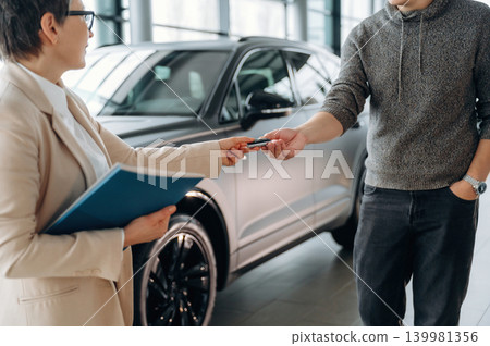 Businesswoman handing car keys to customer in a modern showroom, showcasing the vehicle purchase process and customer service experience Businesswoman handing car keys to customer in a modern showroom, showcasing the vehicle purchase process and customer service experience 139981356
