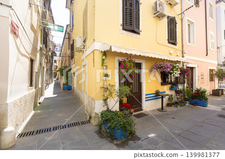 A narrow street is lined with colorful buildings, with potted plants and flowers adorning the entrances in Piran, Slovenia 139981377