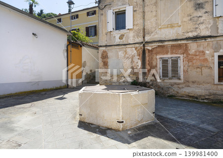 A stone well sits in the middle of a sun-drenched courtyard between weathered buildings. The scene is an outdoor view of a narrow street of Piran, Slovenia A stone well sits in the middle of a sun-drenched courtyard between weathered buildings. The scene is an outdoor view of a narrow street of Piran, Slovenia 139981400