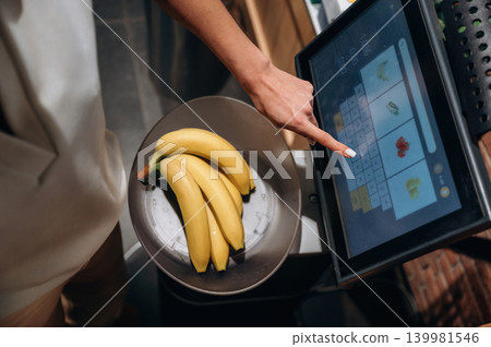 Woman weighing fresh bananas on digital scale in grocery store, interacting with touchscreen display, showcasing modern shopping technology and healthy choices Woman weighing fresh bananas on digital scale in grocery store, interacting with touchscreen display, showcasing modern shopping technology and healthy choices 139981546