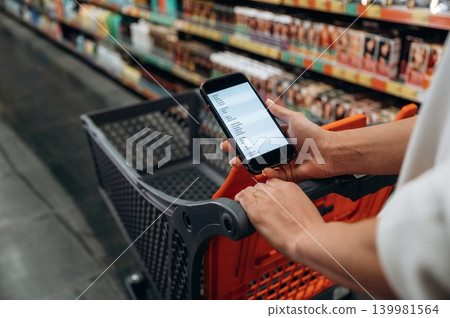 Person holding smartphone while shopping in grocery store, with shopping cart in foreground and shelves filled with products in the background, showcasing modern shopping experience 139981564
