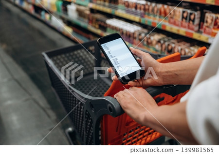 Individual holding smartphone while shopping in a grocery store, with shopping cart and colorful product shelves in the background, showcasing modern shopping experience 139981565