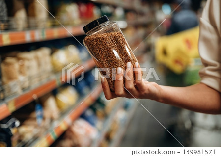 Hand holding a transparent jar filled with grains in a grocery store aisle, surrounded by colorful packaging and shelves of various food products Hand holding a transparent jar filled with grains in a grocery store aisle, surrounded by colorful packaging and shelves of various food products 139981571