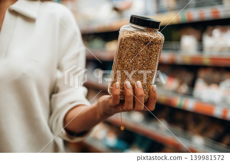 Woman holding a transparent jar filled with grains in a grocery store aisle, showcasing healthy food choices and sustainable shopping habits Woman holding a transparent jar filled with grains in a grocery store aisle, showcasing healthy food choices and sustainable shopping habits 139981572