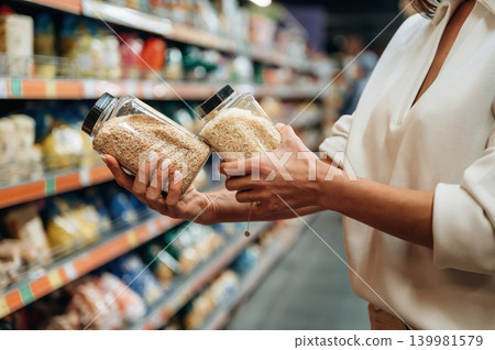 Woman comparing two jars of grains in a grocery store aisle, surrounded by colorful packaging, showcasing the decision-making process in food selection Woman comparing two jars of grains in a grocery store aisle, surrounded by colorful packaging, showcasing the decision-making process in food selection 139981579