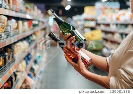 Individual is selecting between two bottles of sparkling wine in a grocery store aisle, showcasing the decision-making process in beverage shopping Individual is selecting between two bottles of sparkling wine in a grocery store aisle, showcasing the decision-making process in beverage shopping 139981586