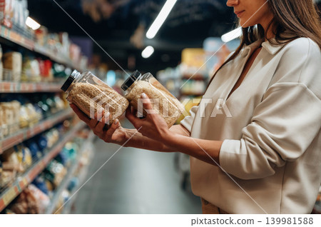 Woman comparing two jars of grains in a grocery store aisle, surrounded by shelves filled with various food products, showcasing shopping choices and healthy living Woman comparing two jars of grains in a grocery store aisle, surrounded by shelves filled with various food products, showcasing shopping choices and healthy living 139981588