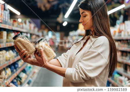 Woman comparing two jars of grains in a grocery store aisle, surrounded by shelves filled with various food products, showcasing shopping choices and healthy options Woman comparing two jars of grains in a grocery store aisle, surrounded by shelves filled with various food products, showcasing shopping choices and healthy options 139981589