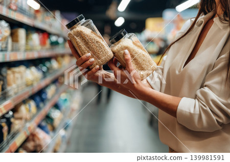 Woman comparing two jars of grains in a grocery store aisle, showcasing healthy food choices and the importance of nutrition in daily life Woman comparing two jars of grains in a grocery store aisle, showcasing healthy food choices and the importance of nutrition in daily life 139981591