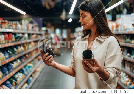 Woman in casual attire is comparing two jars of food products while shopping in a grocery store aisle, surrounded by colorful shelves filled with various items Woman in casual attire is comparing two jars of food products while shopping in a grocery store aisle, surrounded by colorful shelves filled with various items 139981593