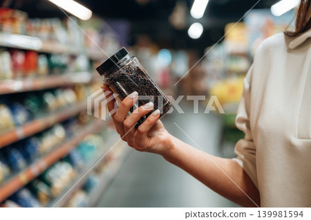 Woman holding a jar of loose tea leaves in a grocery store aisle, surrounded by colorful packaging, showcasing the variety of products available for tea enthusiasts 139981594