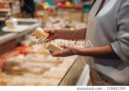 Woman selecting cheese in a grocery store, holding two different types of cheese, showcasing the variety and choices available in the dairy section Woman selecting cheese in a grocery store, holding two different types of cheese, showcasing the variety and choices available in the dairy section 139981595