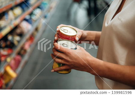 Woman holding two cans in a grocery store aisle, surrounded by colorful products on shelves, showcasing shopping experience and decision-making process Woman holding two cans in a grocery store aisle, surrounded by colorful products on shelves, showcasing shopping experience and decision-making process 139981596