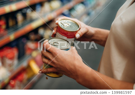 Woman holding two cans while shopping in a grocery store aisle, surrounded by colorful product shelves, showcasing decision-making in food selection Woman holding two cans while shopping in a grocery store aisle, surrounded by colorful product shelves, showcasing decision-making in food selection 139981597