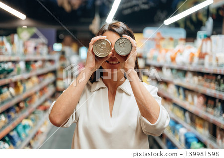 Woman in grocery store playfully holding cans in front of her eyes, surrounded by colorful shelves filled with various products, showcasing a fun shopping experience Woman in grocery store playfully holding cans in front of her eyes, surrounded by colorful shelves filled with various products, showcasing a fun shopping experience 139981598