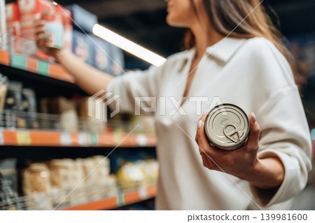 Woman shopping for groceries in a supermarket, holding a can while reaching for another item on the shelf, surrounded by various food products and packaging 139981600