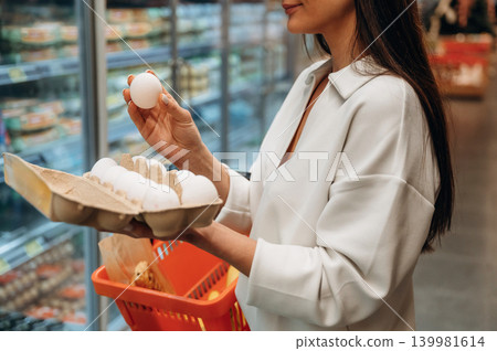 Woman in white blouse is selecting eggs from a carton while shopping in a grocery store, showcasing the food selection process and consumer choices Woman in white blouse is selecting eggs from a carton while shopping in a grocery store, showcasing the food selection process and consumer choices 139981614