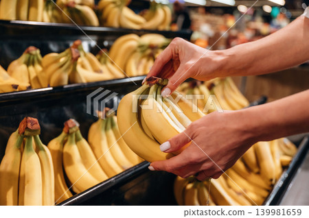 Hands selecting ripe bananas from a display in a grocery store, showcasing fresh produce and vibrant colors in a bustling market environment Hands selecting ripe bananas from a display in a grocery store, showcasing fresh produce and vibrant colors in a bustling market environment 139981659