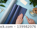 Hands cutting blue polka dot wrapping paper with a craft knife. Ribbon, scissors, gift tag, and pearl decorations on a light blue background. 139982076