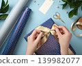 Hands wrapping a gift box with satin ribbon on blue background. Flat lay with wrapping paper, scissors, and tag 139982077