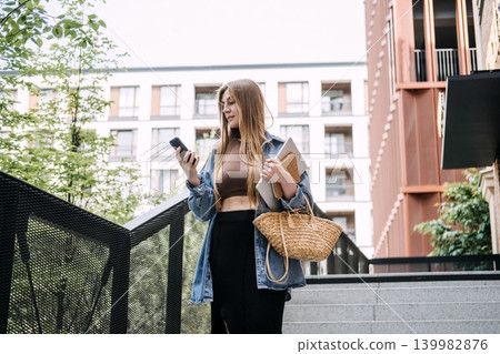 Young woman stands on city stairs looking at smartphone while holding laptop. On the fly decision making, active professional lifestyle, business mobility trends. 139982876