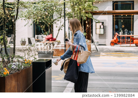 Young woman throws disposable cup into public trash bin on city street. Proper waste management, urban litter control, responsible rubbish disposal. Young woman throws disposable cup into public trash bin on city street. Proper waste management, urban litter control, responsible rubbish disposal. 139982884