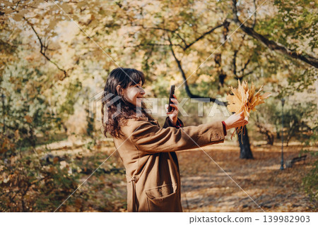 Person takes picture of yellow autumn leaves with smartphone in sunny park. Taking photo with smartphone, mobile photography outdoor, female content creator. Person takes picture of yellow autumn leaves with smartphone in sunny park. Taking photo with smartphone, mobile photography outdoor, female content creator. 139982903
