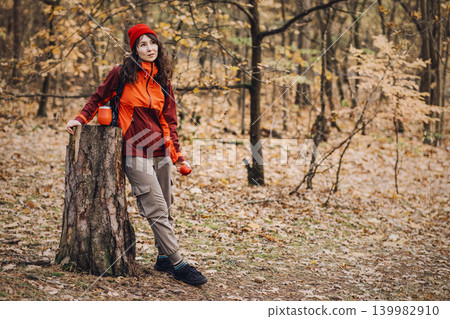 Woman stands by tree stump in autumn forest holding apple and thermal cup. nature therapy experience, forest bathing wellness, mindful outdoor activity, stress relief nature. Woman stands by tree stump in autumn forest holding apple and thermal cup. nature therapy experience, forest bathing wellness, mindful outdoor activity, stress relief nature. 139982910