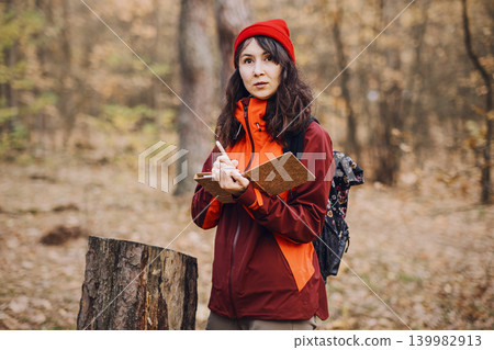 Middle aged woman writes in notebook while standing in autumn forest next to tree stump. Eco-therapy practice, emotional balance outdoors, stress reduction nature. Middle aged woman writes in notebook while standing in autumn forest next to tree stump. Eco-therapy practice, emotional balance outdoors, stress reduction nature. 139982913