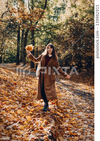 Woman walks along foliage covered path in autumn park holding bunch of leaves. Seasonal Reset Walk, Autumn Emotional Release, Mindful Solitude in Nature. 139982915