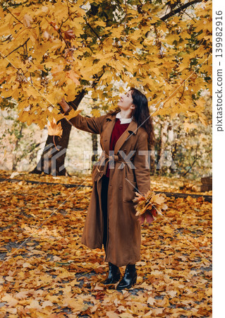 Woman stands in park touching tree branches and holding bunch of fallen leaves. Microadventure local park autumn, Autumn Micro adventures. 139982916