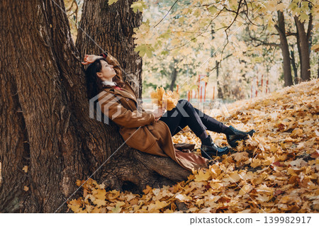 Woman rests against large tree trunk in autumn forest holding leaves with closed eyes. Grounding in nature autumn, Sensory Walking, Physical Earthing, Cycle of Letting Go. 139982917