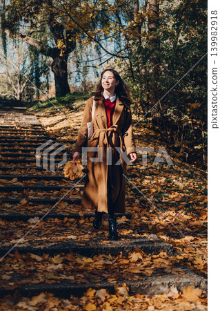 Woman walks down stone steps in autumn park holding leaves. Celebrating autumn seasonal changes, optimism and well-being, authentic happy expressions. 139982918