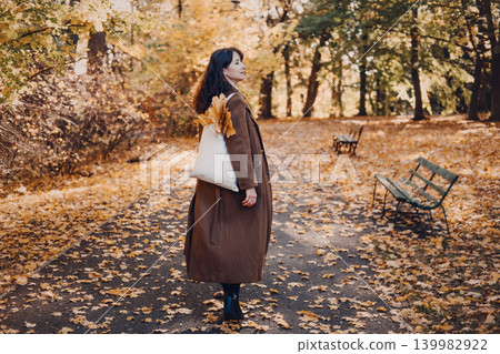Woman stands on path in autumn park holding bag filled with leaves. Sensory autumn experience, mindfulness in changing seasons, living the moment concept. 139982922