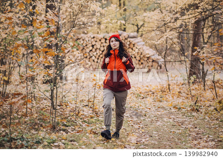 Woman hiker walks through autumn forest with stack of timber logs in background. independent woman hiker, outdoor navigation skills, active leisure security. Woman hiker walks through autumn forest with stack of timber logs in background. independent woman hiker, outdoor navigation skills, active leisure security. 139982940