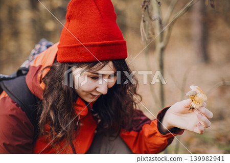 Woman squats in forest holding small wild mushroom for examination. Mushroom identification guide, amateur mycology study, botanical field research, fungal species recognition. Woman squats in forest holding small wild mushroom for examination. Mushroom identification guide, amateur mycology study, botanical field research, fungal species recognition. 139982941