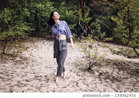 Barefoot woman walks on sand through forest with eyes closed. Mindful living concept, finding inner peace, psychological wellbeing outdoors. Barefoot woman walks on sand through forest with eyes closed. Mindful living concept, finding inner peace, psychological wellbeing outdoors. 139982958