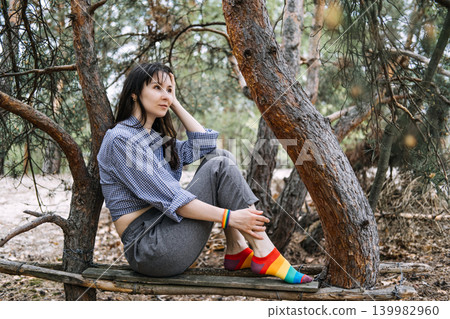 Adult person sits on wooden branch between pine trees in forest. lgbtq mental health, queer mindfulness nature, finding inner peace, self-care for queer. Adult person sits on wooden branch between pine trees in forest. lgbtq mental health, queer mindfulness nature, finding inner peace, self-care for queer. 139982960
