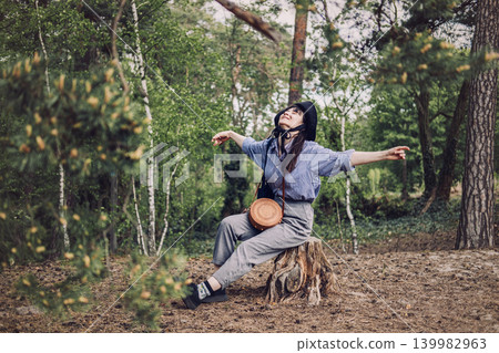 Person wearing hat sits on tree stump in forest with arms outstretched and eyes closed. Nature therapy for stress relief, forest bathing concept, mental well-being in nature. 139982963