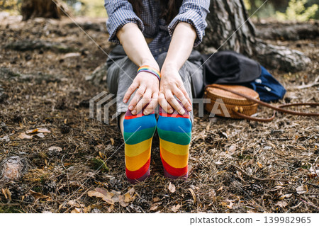 Person sits on forest floor wearing rainbow socks with feet pointed toward camera. LGBTQ representation, authentic queer life, Non-binary person relaxing, pride month accessories. 139982965