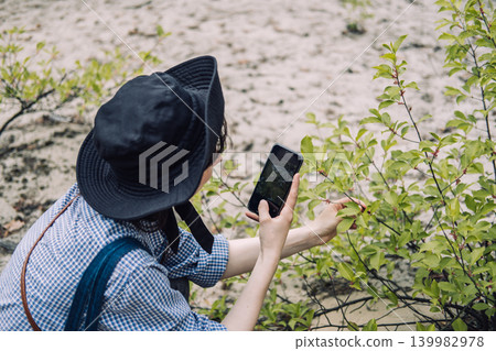 Young woman takes close up photo of green leaves with smartphone in sandy forest. Social media influencer outdoors, capturing nature moments, modern hiker lifestyle. Young woman takes close up photo of green leaves with smartphone in sandy forest. Social media influencer outdoors, capturing nature moments, modern hiker lifestyle. 139982978