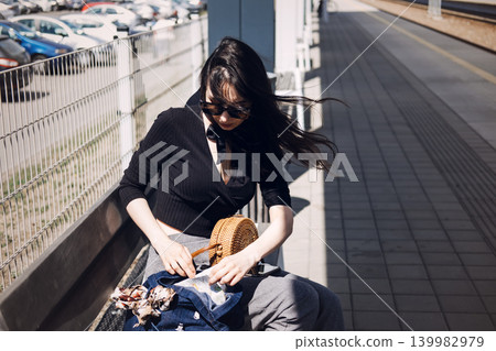 Woman sitting on bench at railway platform looks through bag. Summer vacation solo, millennial travel trends, adventurous lifestyle 139982979