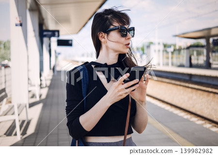 Woman stands on railway platform while looking at smartphone. Slow travel movement, analog navigation, conscious tourism experience Woman stands on railway platform while looking at smartphone. Slow travel movement, analog navigation, conscious tourism experience 139982982