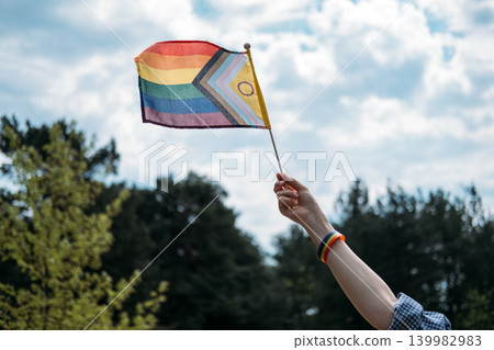 Hand holding intersex inclusive progress pride flag waves against cloudy sky. Symbolic LGBTQ accessories, celebrating identity outdoors, summer pride events. Hand holding intersex inclusive progress pride flag waves against cloudy sky. Symbolic LGBTQ accessories, celebrating identity outdoors, summer pride events. 139982983