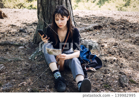 Person sits on ground using smartphone and paper map in forest. Hiking trail navigation, outdoor tourism activity, wilderness exploration trip. Person sits on ground using smartphone and paper map in forest. Hiking trail navigation, outdoor tourism activity, wilderness exploration trip. 139982995