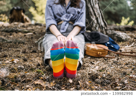 Person sits on forest ground with hands on feet wearing rainbow socks. authentic self representation, quiet pride moment, everyday lgbt life. Person sits on forest ground with hands on feet wearing rainbow socks. authentic self representation, quiet pride moment, everyday lgbt life. 139982998