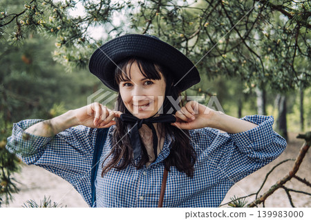 Person wearing hat stands under pine tree branches in forest and adjusts bow on collar while looking at camera. Natural fiber textiles, plastic-free living advocate, slow fashion choices. Person wearing hat stands under pine tree branches in forest and adjusts bow on collar while looking at camera. Natural fiber textiles, plastic-free living advocate, slow fashion choices. 139983000