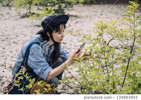 Woman crouches while using smartphone to identify green plant in sandy forest area. Smartphone photography hobby, mobile app for plants, digital content creation. Woman crouches while using smartphone to identify green plant in sandy forest area. Smartphone photography hobby, mobile app for plants, digital content creation. 139983003