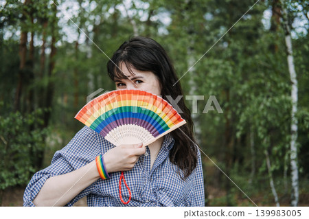 Person holds rainbow fan over face while looking at camera in forest. lgbtq queer identity symbol, coming out support, proud and visible. 139983005